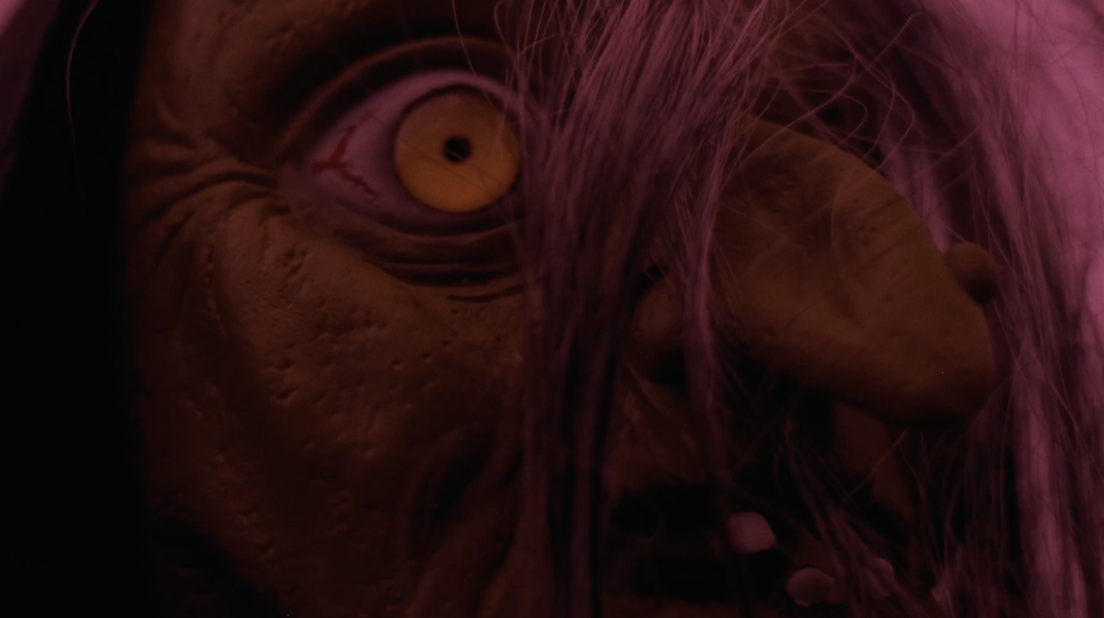 Extreme close-up of the witch's yellow eye behind strands of hair, washed in magenta